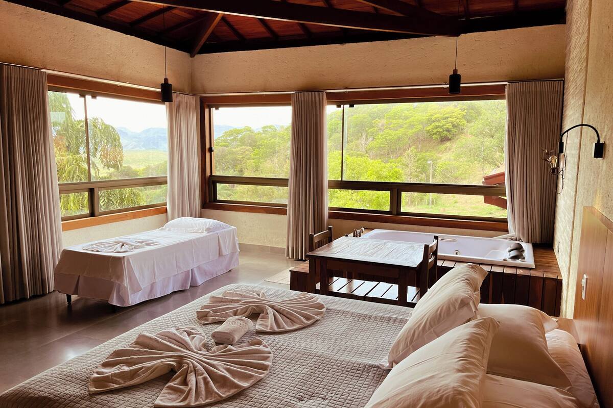 A room with two beds, a table, and a bench. There are large windows with curtains, and the room has a wooden ceiling.
