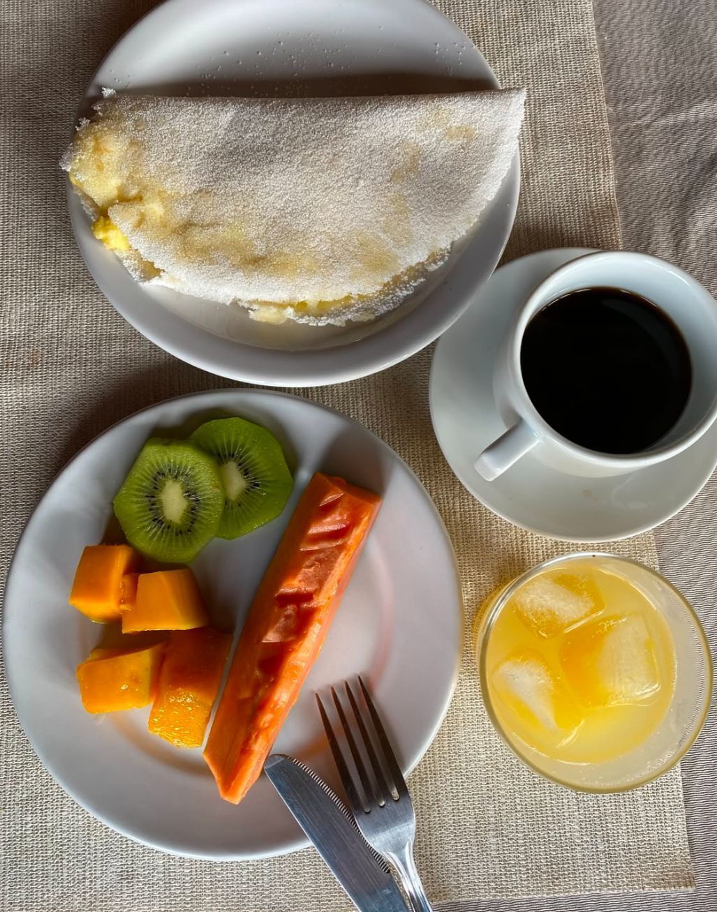 Un plato de desayuno con una porción de pastel, kiwi, mango y salmón, una taza de café y un vaso de jugo de naranja.