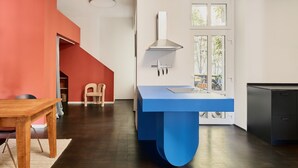 A modern kitchen with a blue island, a dining area with a wooden table, and a staircase leading to an upper level. - Poets Place | Design Apartment in Kreuzberg (Berlin)