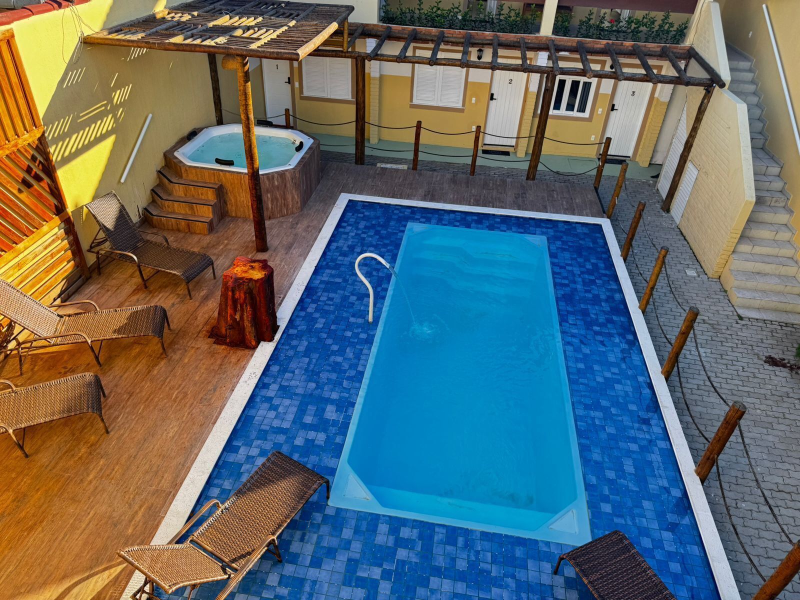 Piscina retangular com esguicho, deck ladrilhado, espreguiçadeiras de madeira ao redor e ofurô.