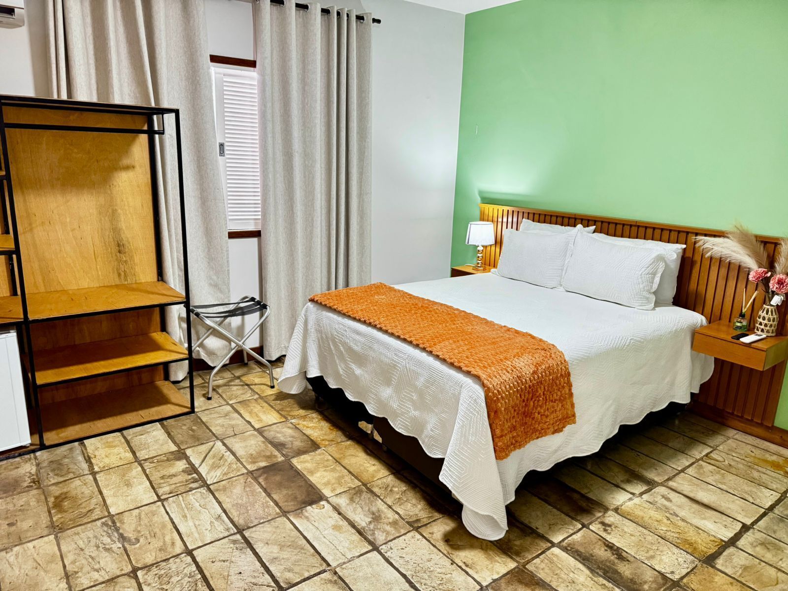 A hotel room with a bed, a wooden shelf unit, a small table with a lamp, and a nightstand.