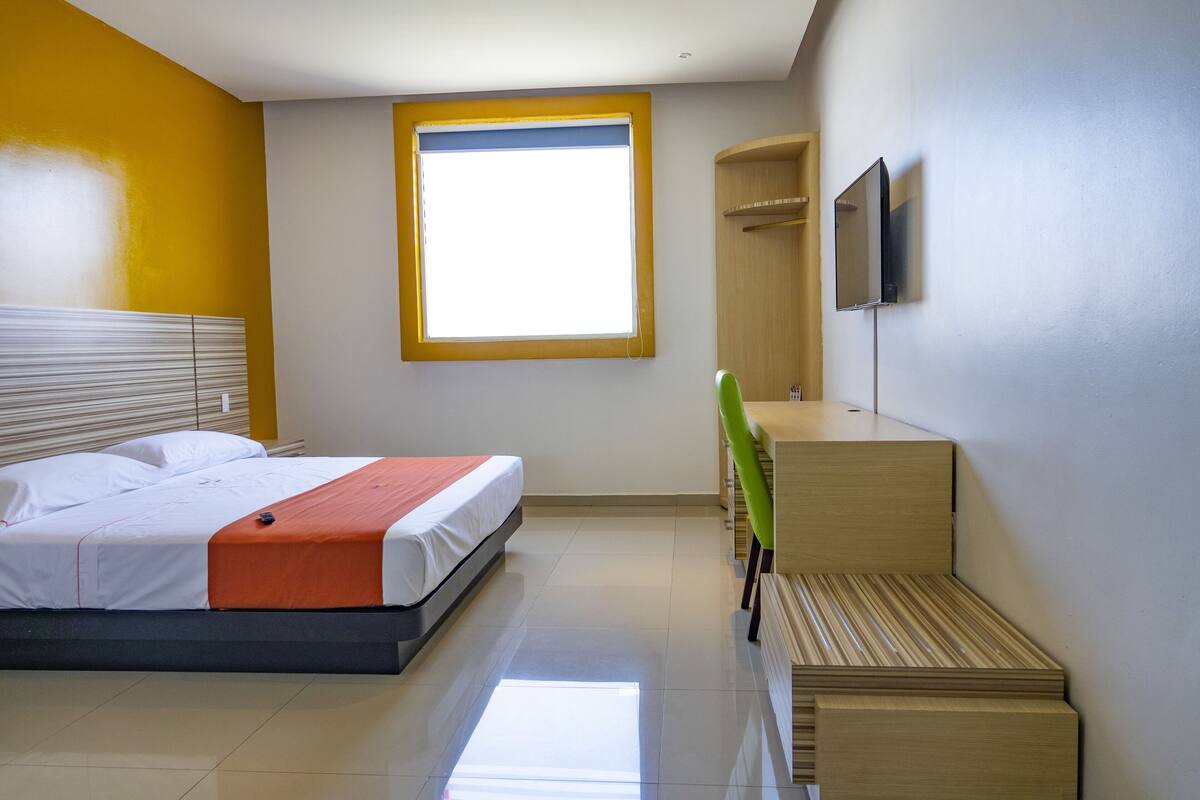 A modern hotel room with a bed, a small desk, a chair, a TV mounted on the wall, and a window with blinds.