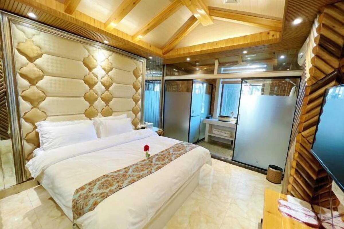 A bedroom with a large bed, a wooden ceiling, a bathroom with a mirror, and a TV.