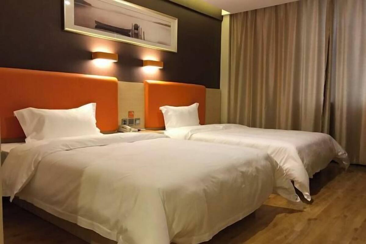 A hotel room with two beds, each with white bedding and orange headboards. There is a framed picture on the wall above the beds and a curtain on the window.