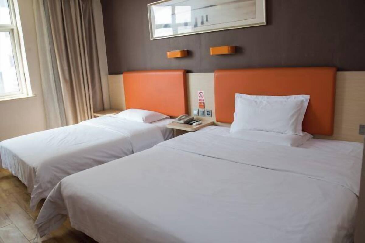 A hotel room with two beds, each with white bedding and orange headboards. There is a window with curtains, a framed picture on the wall, and a bedside table with a phone and a small item on it.