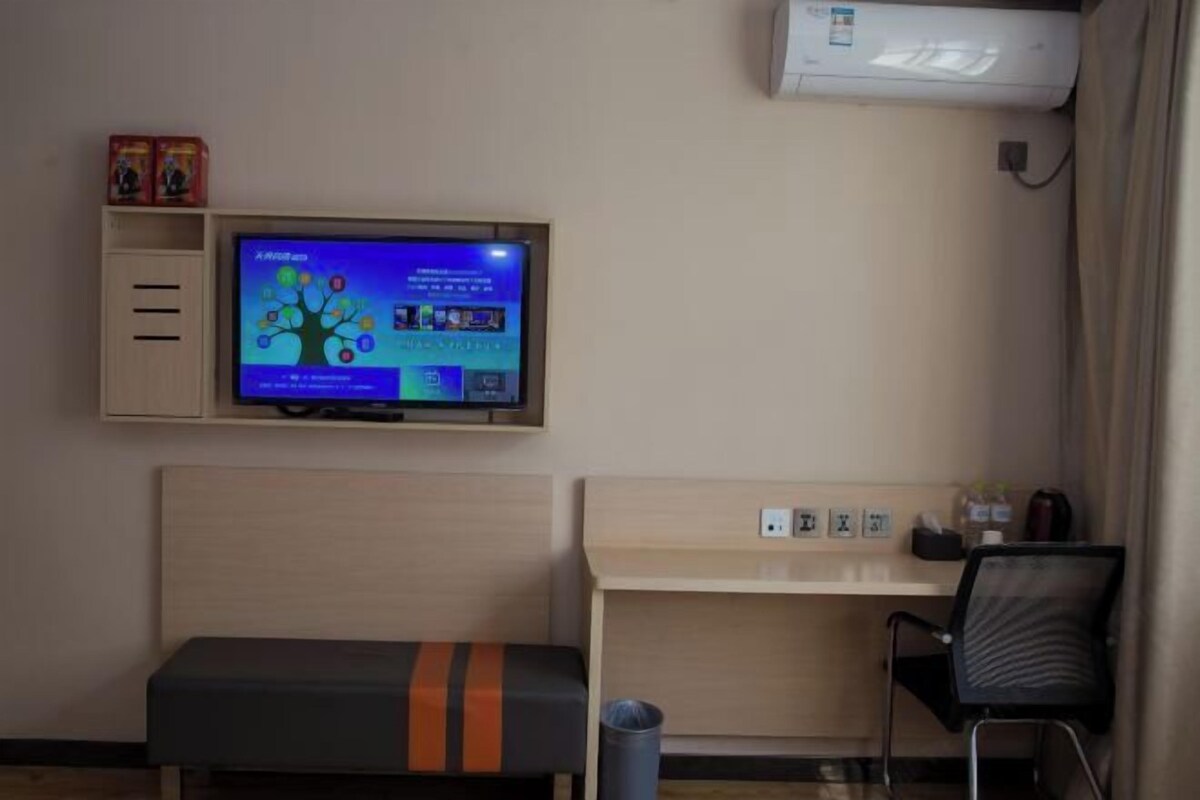 A hotel room with a television mounted on the wall, a desk with a chair, and a bench.