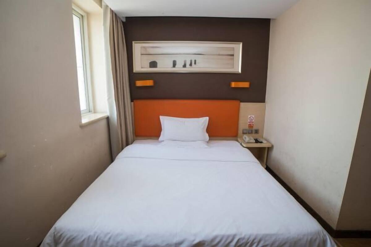 A hotel room with a single bed, a nightstand, a framed picture above the bed, and a window on the left.