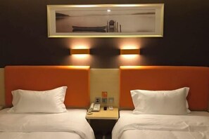 Una camera d'albergo con due letti, ognuno con biancheria bianca e testate arancioni. Sopra i letti c'è un quadro incorniciato, un comodino con un telefono e una piccola lampada, e un punto luce fissato al muro tra i letti.