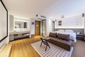 Une chambre d’hôtel moderne équipée d’un lit, d’un canapé, d’une petite table, d’un miroir et d’une salle de bain avec douche.