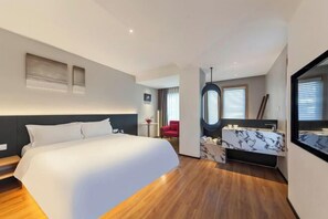 Une chambre d’hôtel moderne avec un grand lit, une coiffeuse avec un miroir et un petit coin salon avec un fauteuil rouge.