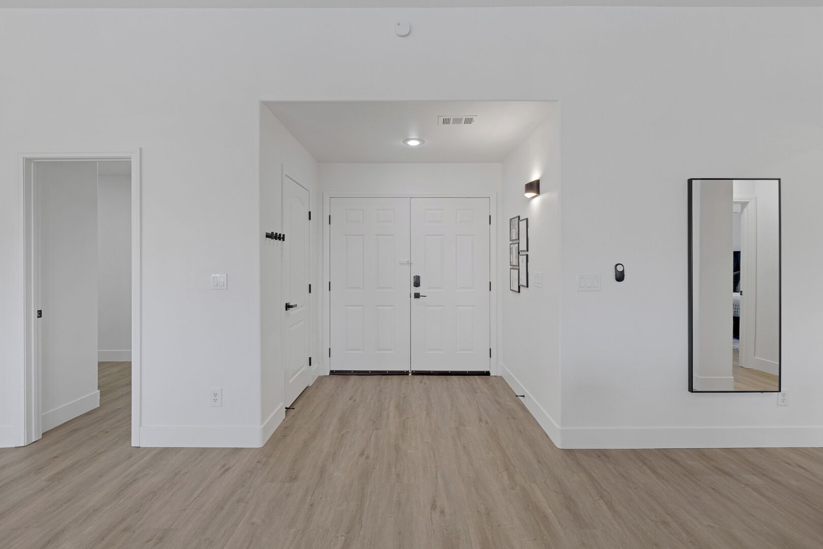Un pasillo con paredes blancas, piso de madera y un espejo grande en la pared derecha. Hay dos puertas blancas al final del pasillo.