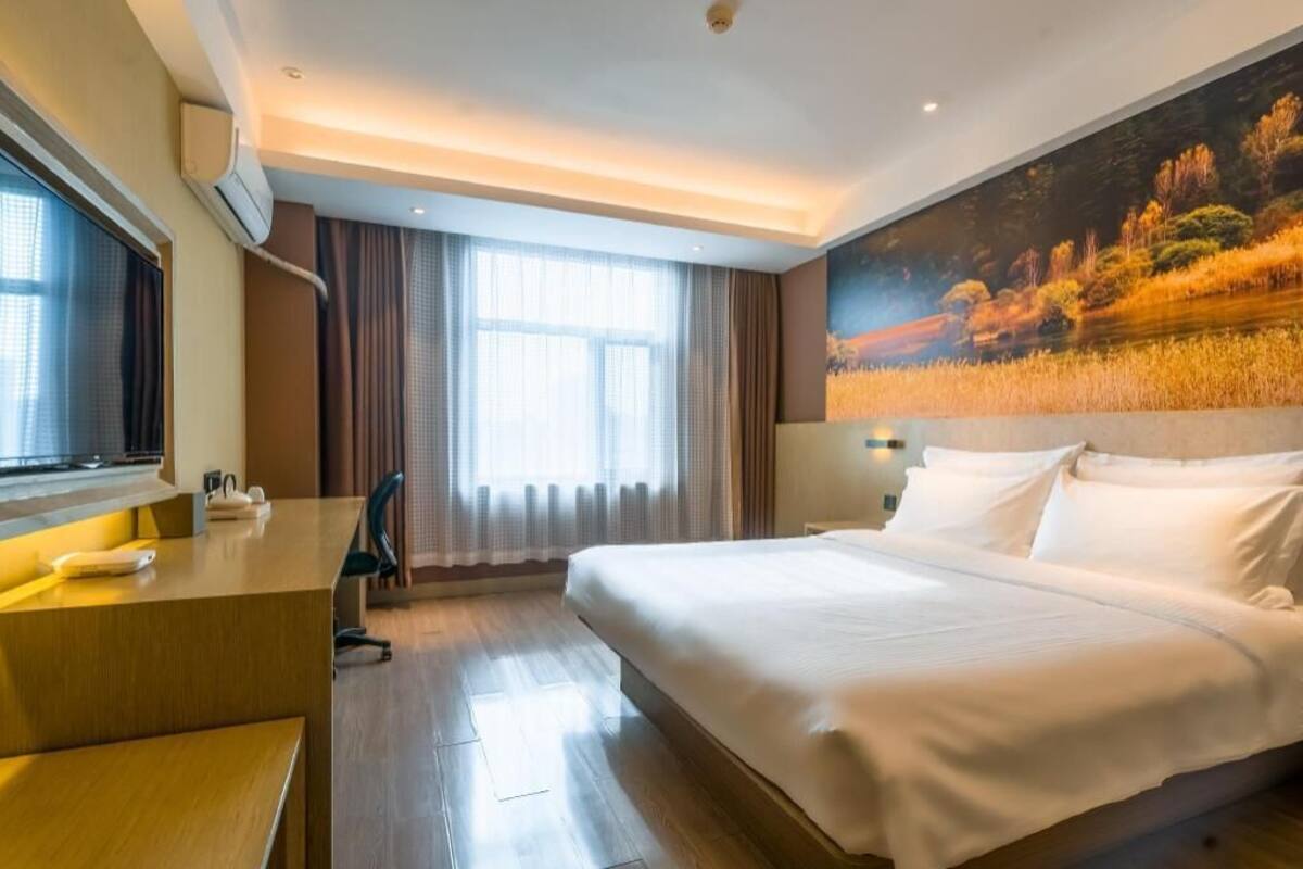 A hotel room with a bed, a desk, a chair, a large window with curtains, and a painting on the wall.