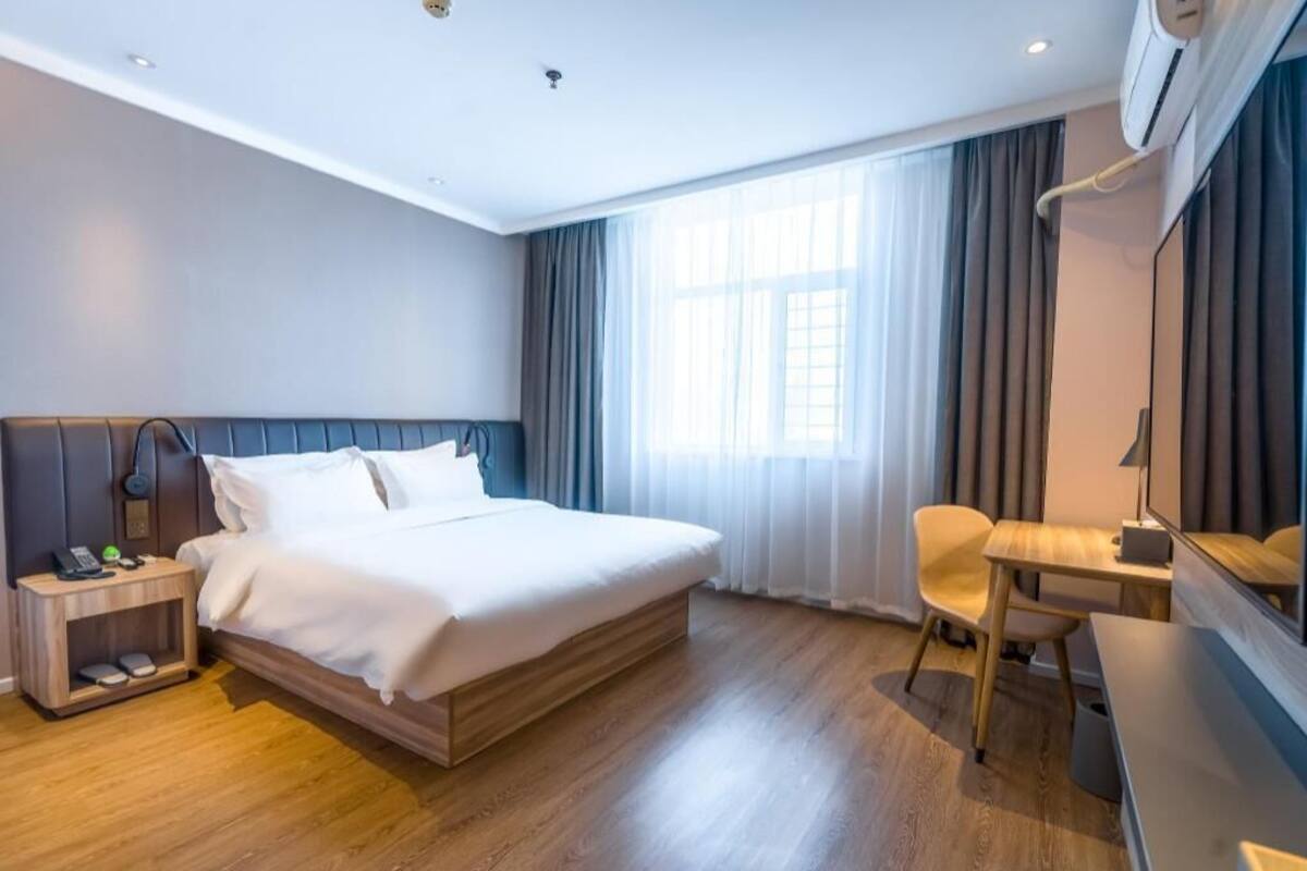 A hotel room with a bed, bedside table, chair, desk, and a large window with curtains.