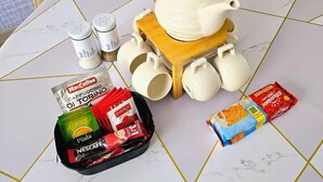 Un tavolo con un bollitore bianco su un supporto in legno, un vaso di tulipani gialli e diversi prodotti per caffè e tè.