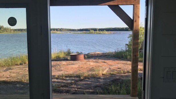 A wooden deck leading to a sandy beach with a body of water and trees in the background. - Tiny lakefront cabin at Loon Straits on Lake Winnipeg. Perfect for couples! (Loon Straits)