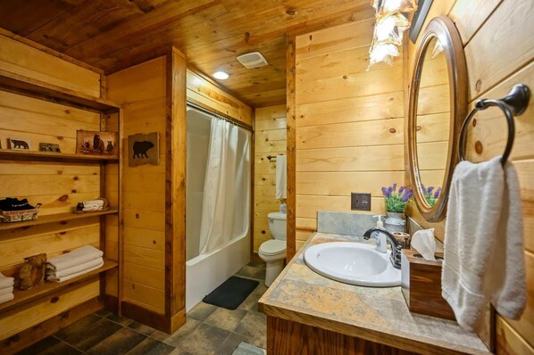 A wooden cabin bathroom with a bathtub, toilet, and sink. Shelves with towels and toiletries are visible. - Whitetail Retreat Cabin for 13! (Broken Bow)