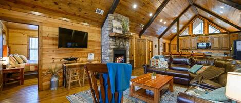 A cozy cabin interior with a stone fireplace, wooden ceiling beams, a leather sofa, and a dining area.