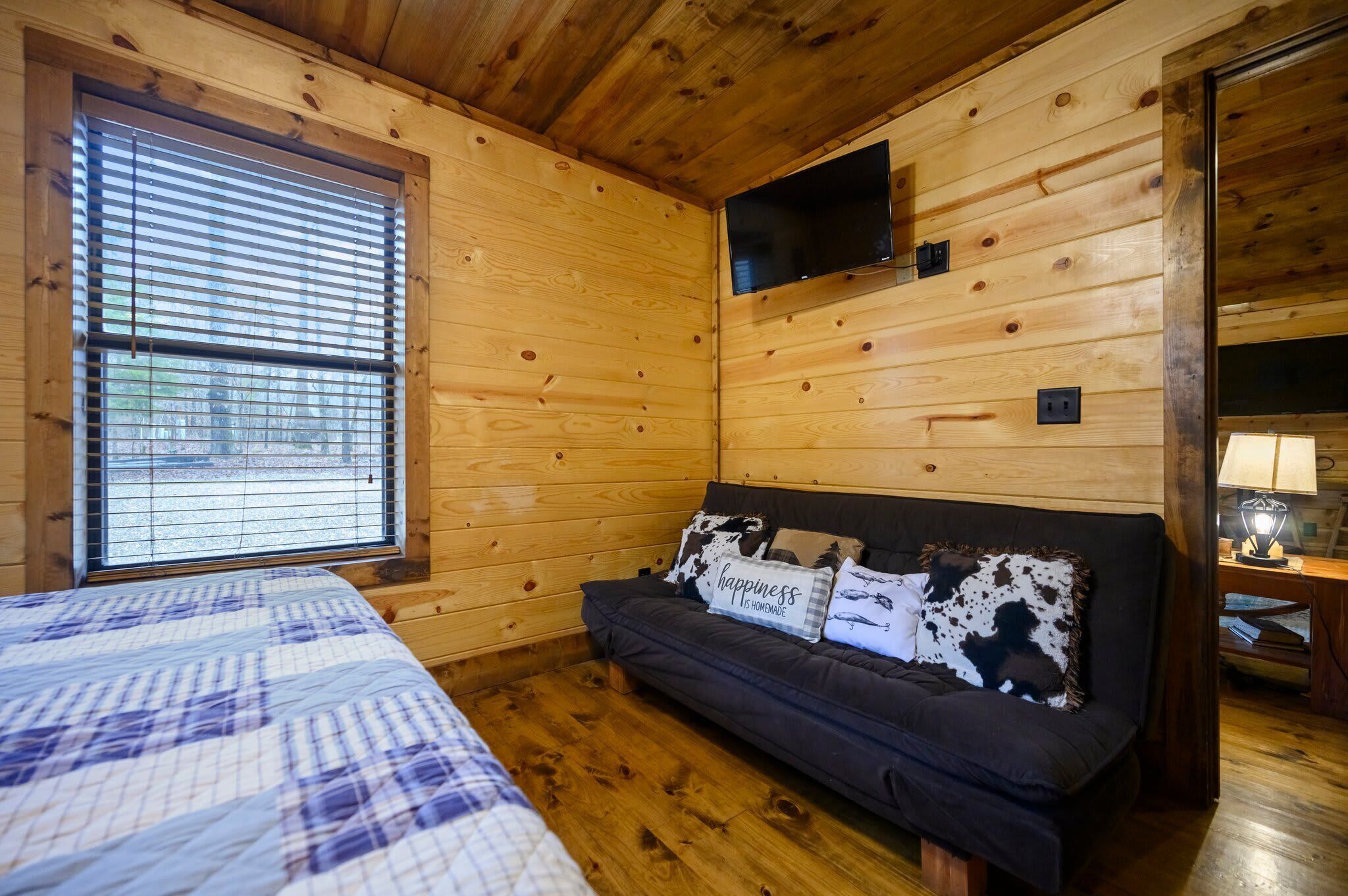A cozy room with wooden walls, a bed with a patterned bedspread, a dark sofa with decorative pillows, and a flat-screen TV mounted above the sofa.
