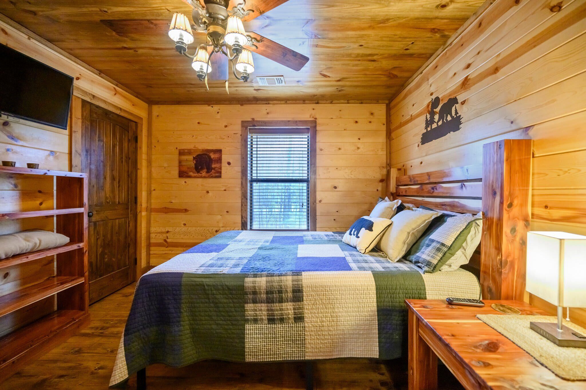 Un dormitorio de cabaña de madera con cama, ventilador de techo, ventana y un mural con silueta de oso.