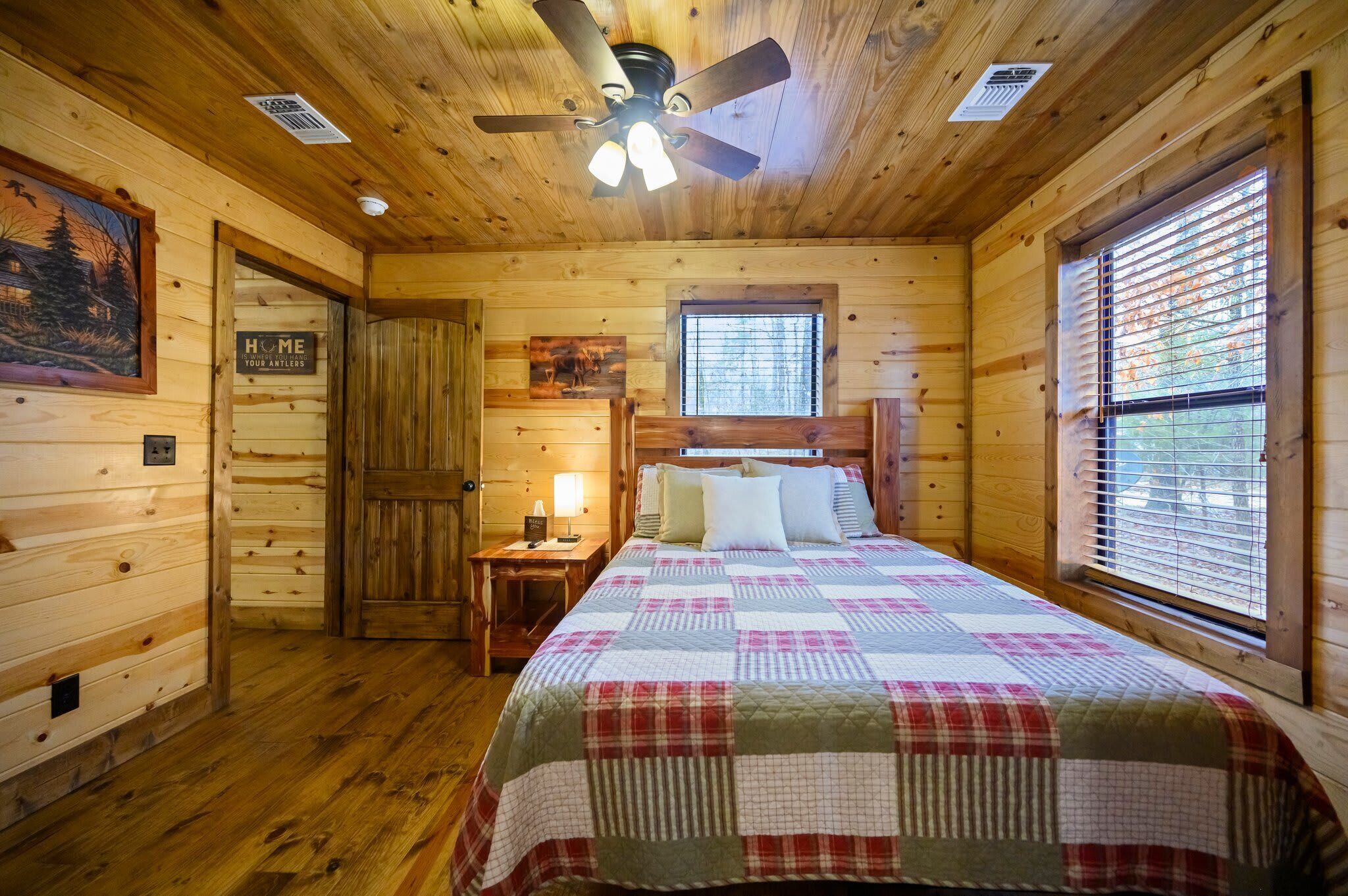 Een slaapkamer in een houten hut met een bed, een nachtkastje, een plafondventilator en een raam met jaloezieën.