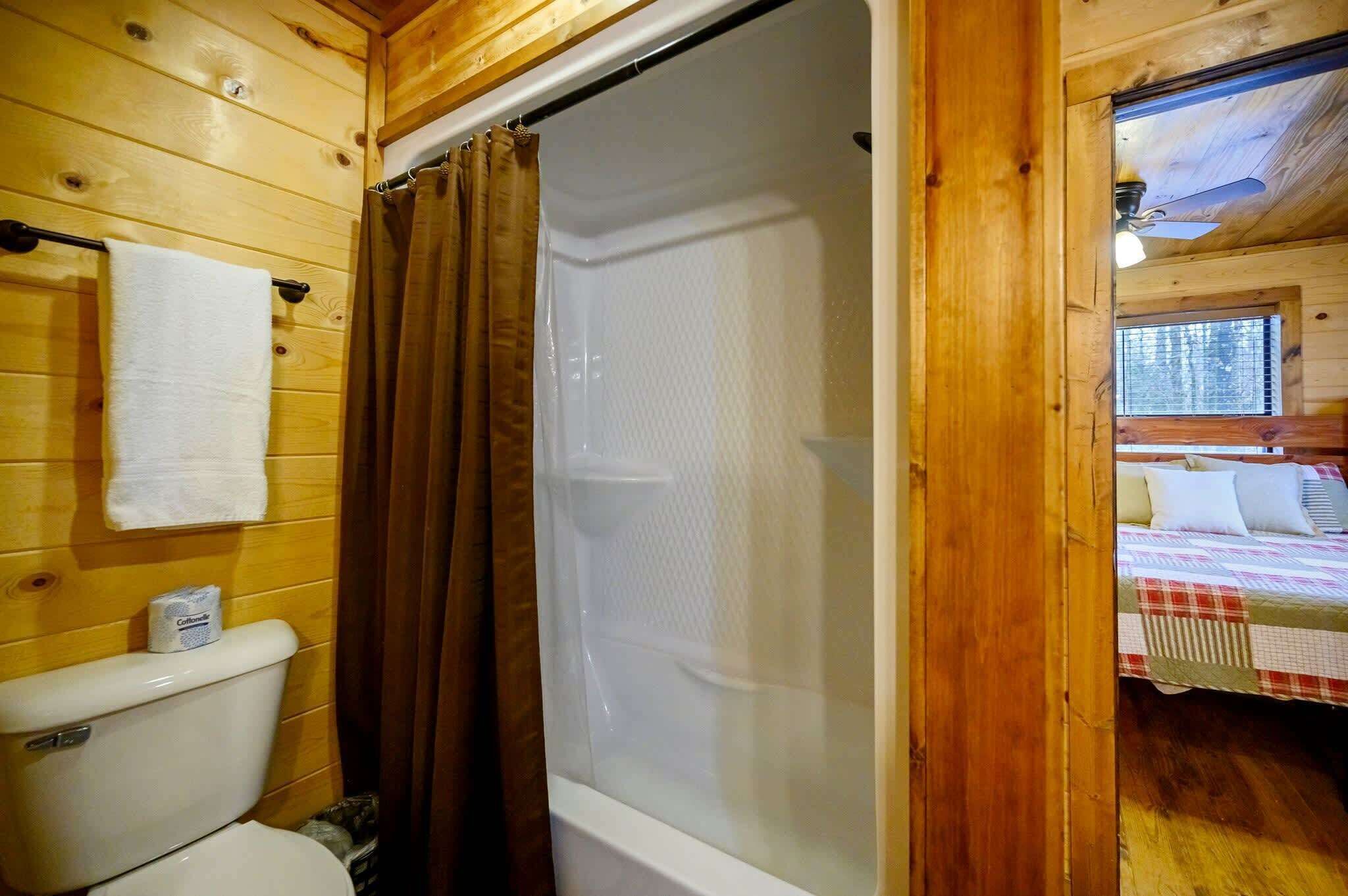 Une salle de bain rustique, aménagée dans une cabane en bois, avec des toilettes, un rideau de douche et un lit recouvert d’une couverture à carreaux.