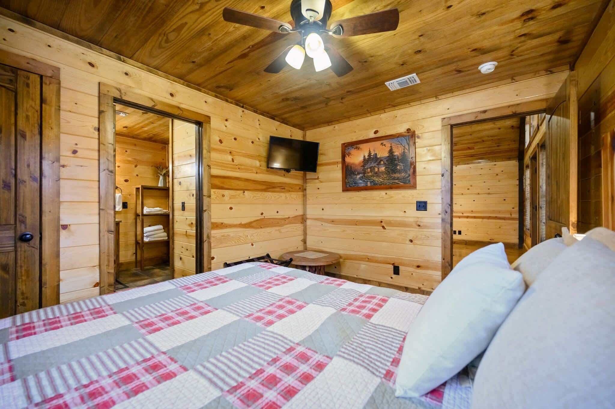 Habitación de cabaña de madera con una cama, un ventilador de techo, un televisor montado y un cuadro en la pared.