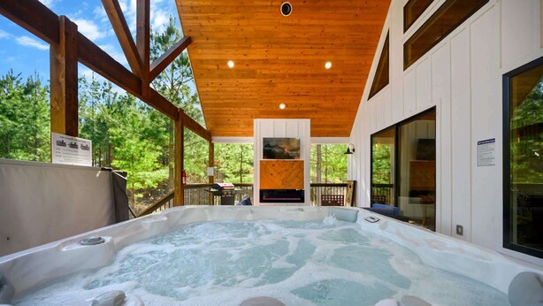 A hot tub in a wooden cabin with large windows and a view of the forest. - Sugar Bear Den Stay for 10! (Broken Bow)