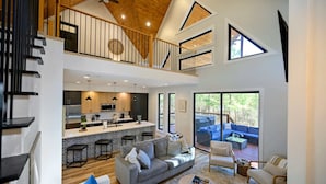 A spacious living area with a high ceiling, a kitchen with a central island, and a balcony with outdoor seating. - Sugar Bear Den Stay for 10! (Broken Bow)