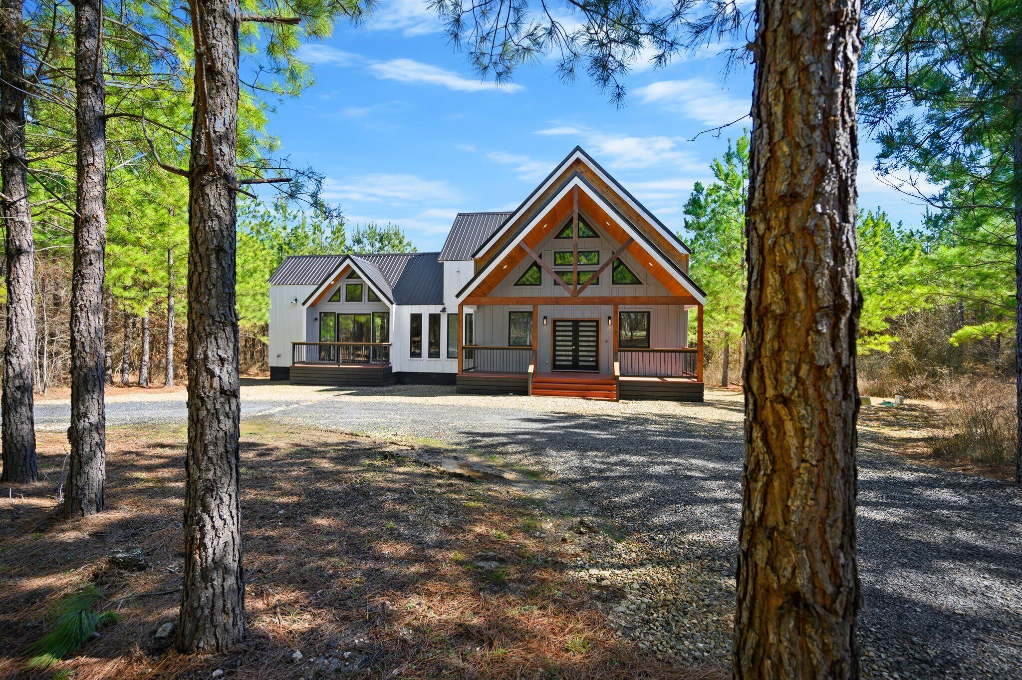 A cabin with a triangular roof, large windows, and a porch. It is surrounded by trees and has a gravel driveway.