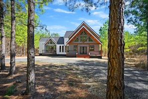 A cabin with a triangular roof, large windows, and a porch. It is surrounded by trees and has a gravel driveway. - Sugar Bear Den Stay for 10! (Broken Bow)