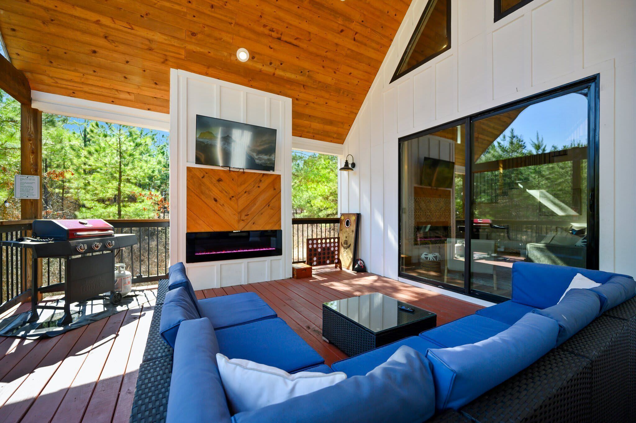 A covered deck with a large sofa, a TV mounted on a wooden wall, and a grill.