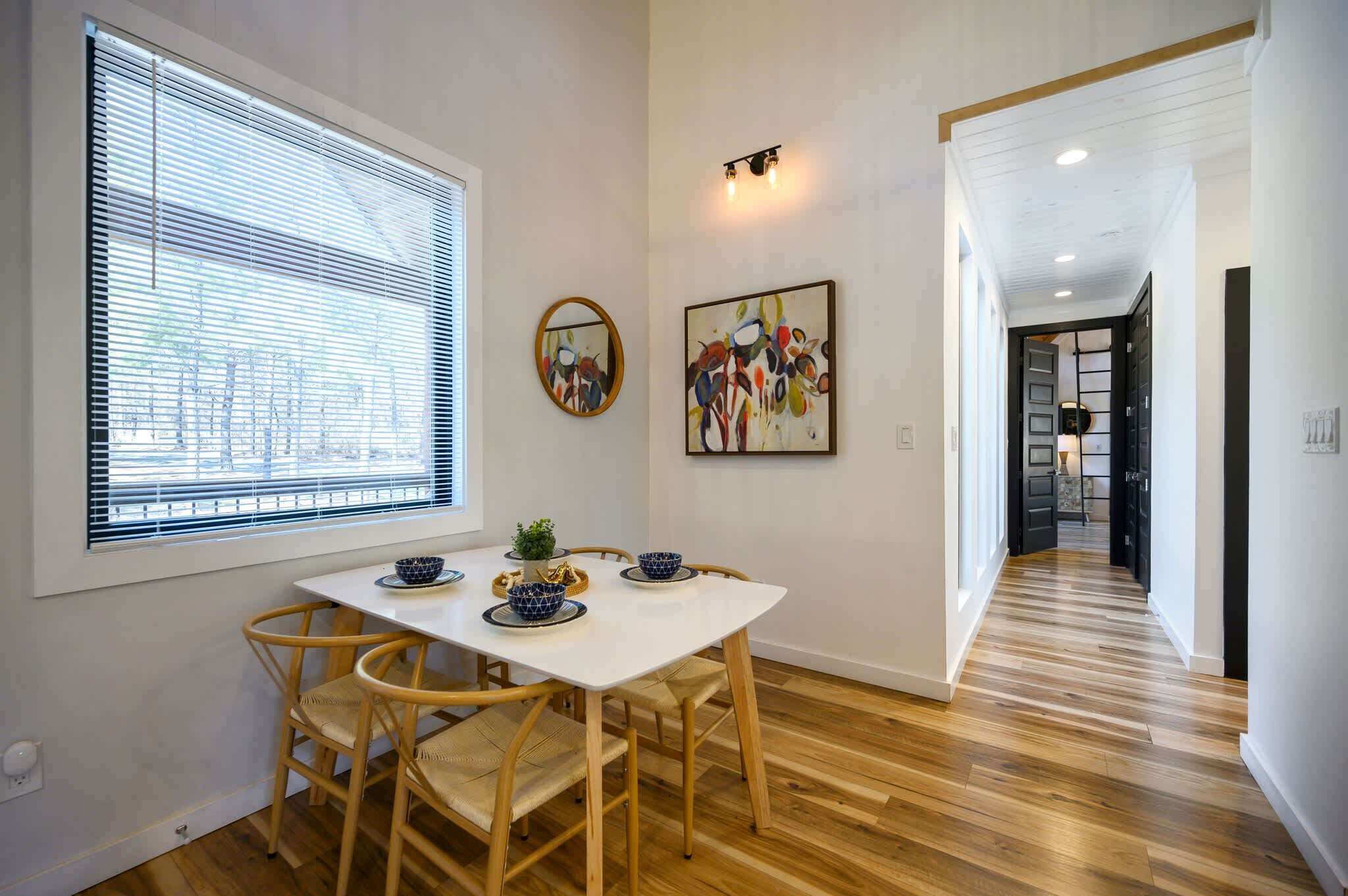Un coin repas avec une table blanche et des chaises en bois, une fenêtre avec des stores et un couloir au plancher en bois.