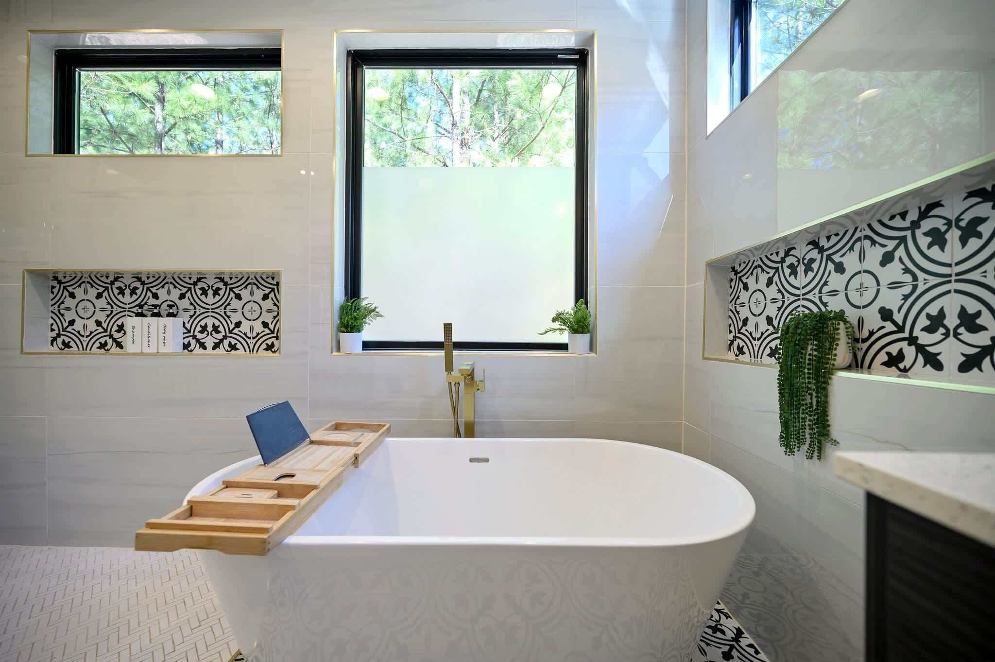 A modern bathroom with a freestanding bathtub, two windows with views of trees, patterned tile walls, and a small plant on the right.