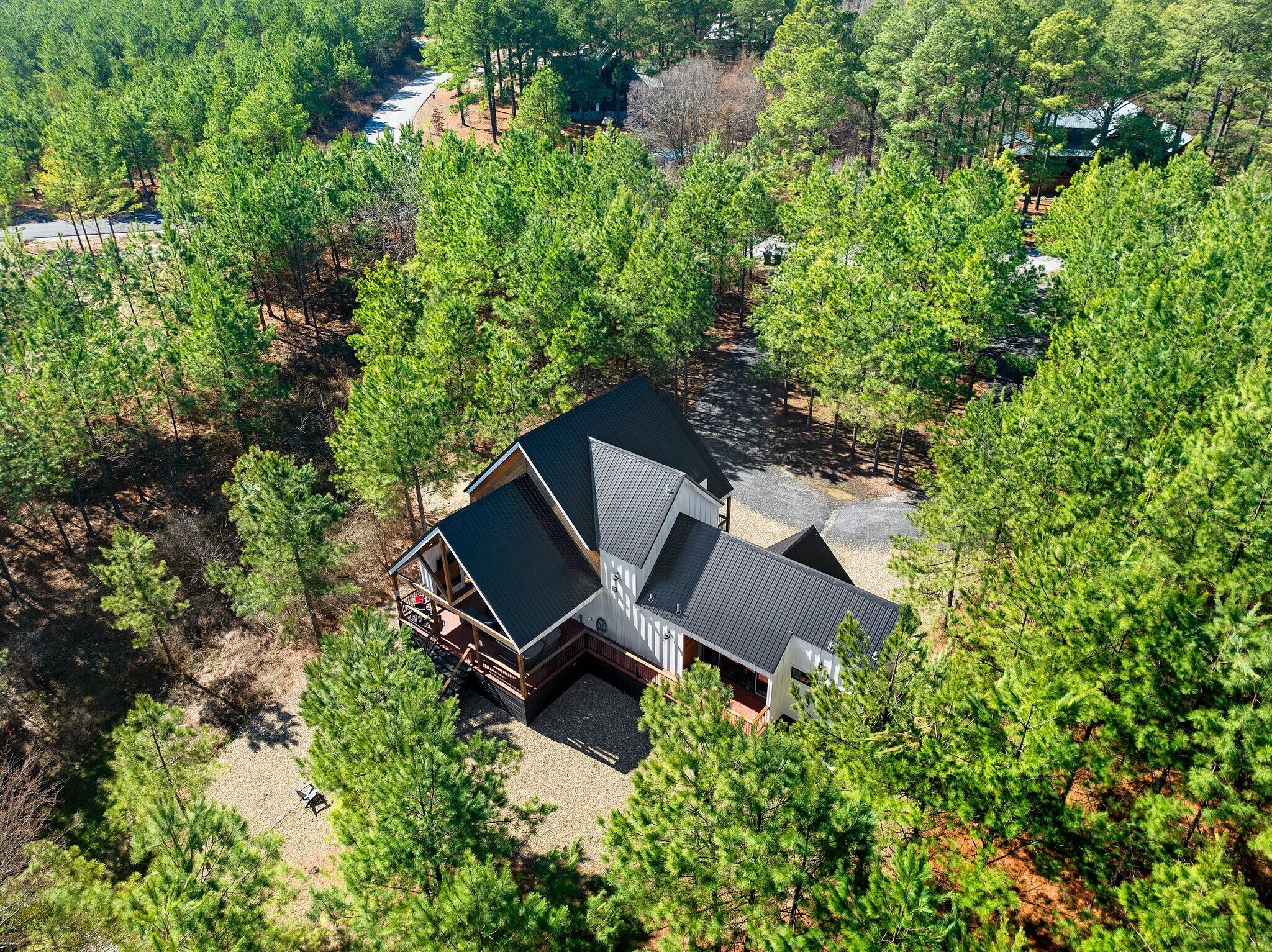 A modern cabin surrounded by dense forest, with a gravel driveway and a wooden deck.