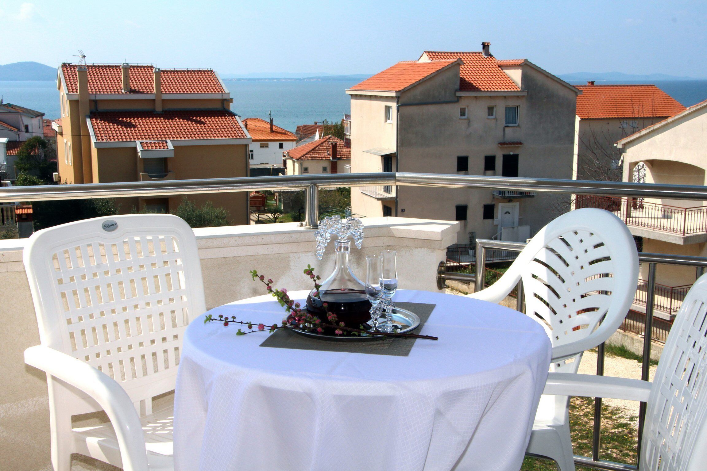 Una mesa redonda con mantel blanco, dos sillas blancas y un vaso de agua en un balcón con vistas a un pueblo costero con edificios de tejados rojos.
