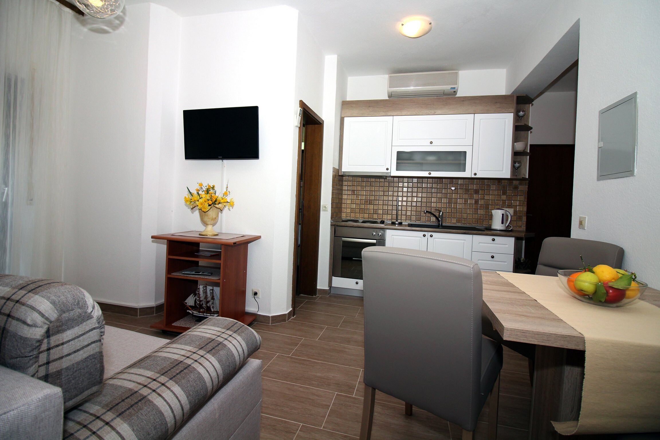 A compact living space with a sofa, a dining table with a fruit bowl, a kitchen area with cabinets and a microwave, and a flat-screen TV mounted on the wall.