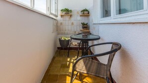 Een klein terras buiten met een ronde tafel en twee rieten stoelen, een plant in een pot op een plank, en een betegelde vloer.