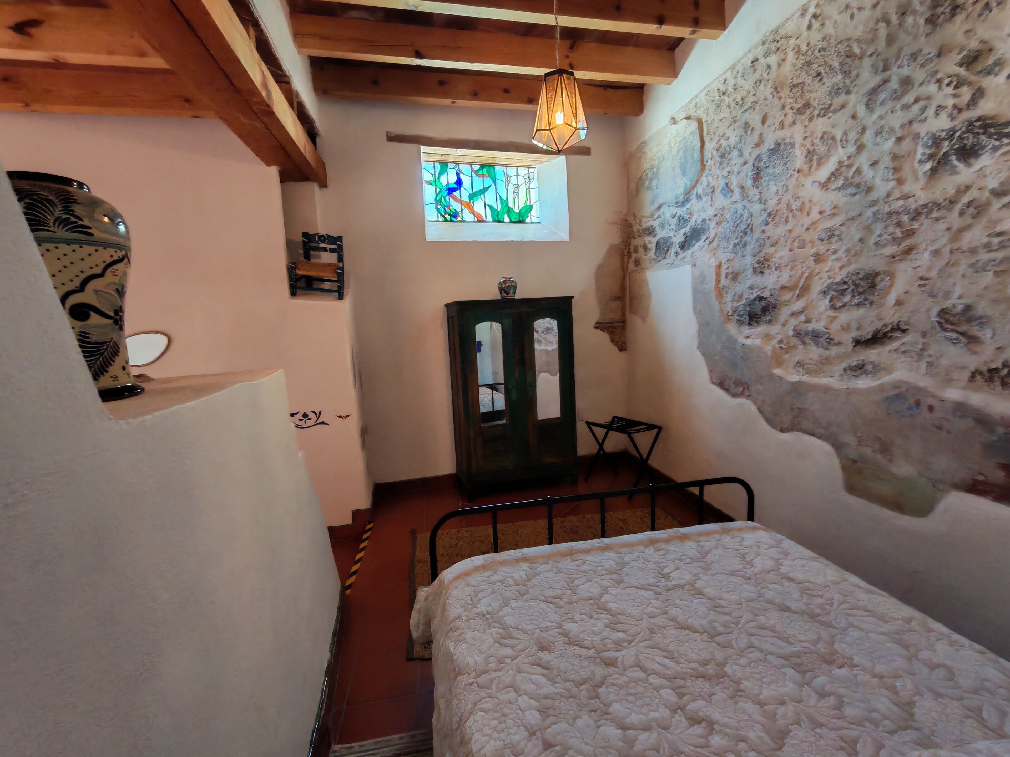 Una stanza piccola con un letto, un soffitto in legno, una parete in pietra, una finestra con vetrata decorata e un armadietto di legno.