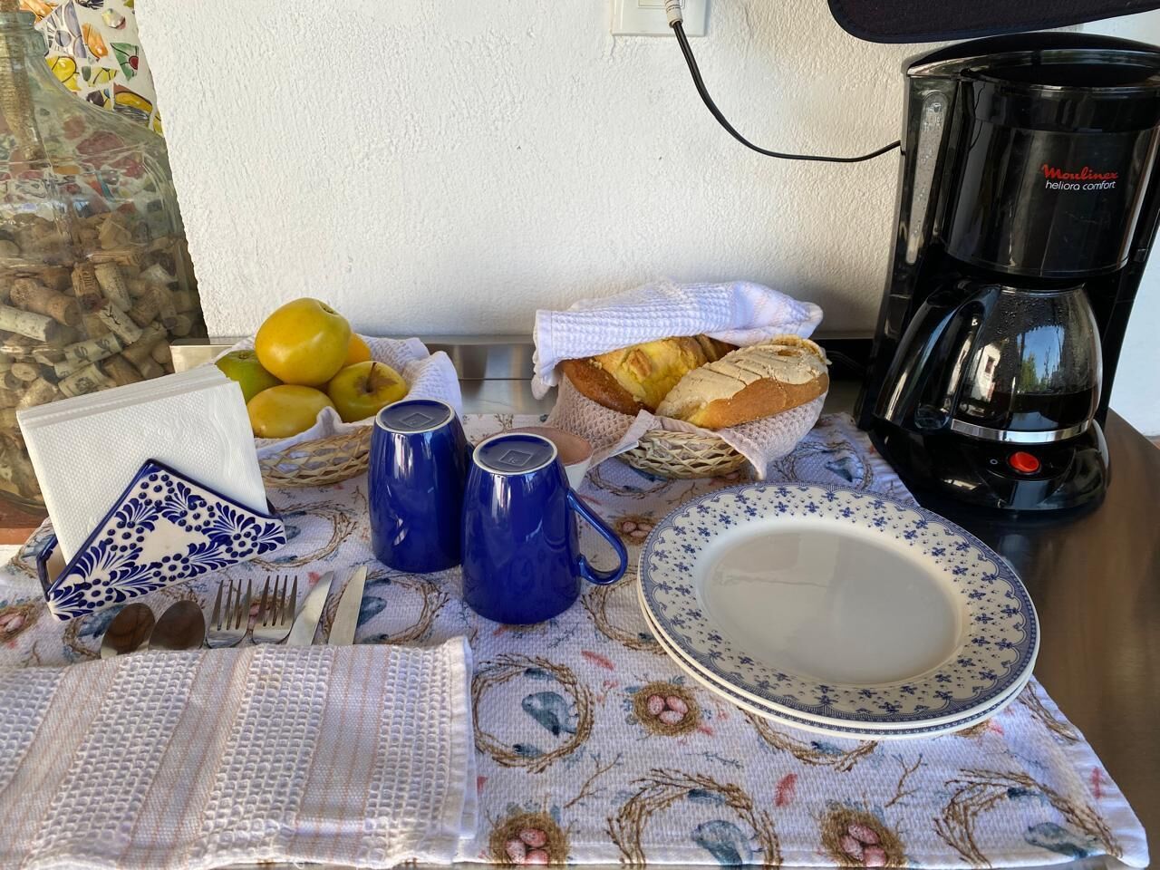 Uma mesa com toalha florida, uma cafeteira, uma cesta de pão e um prato.