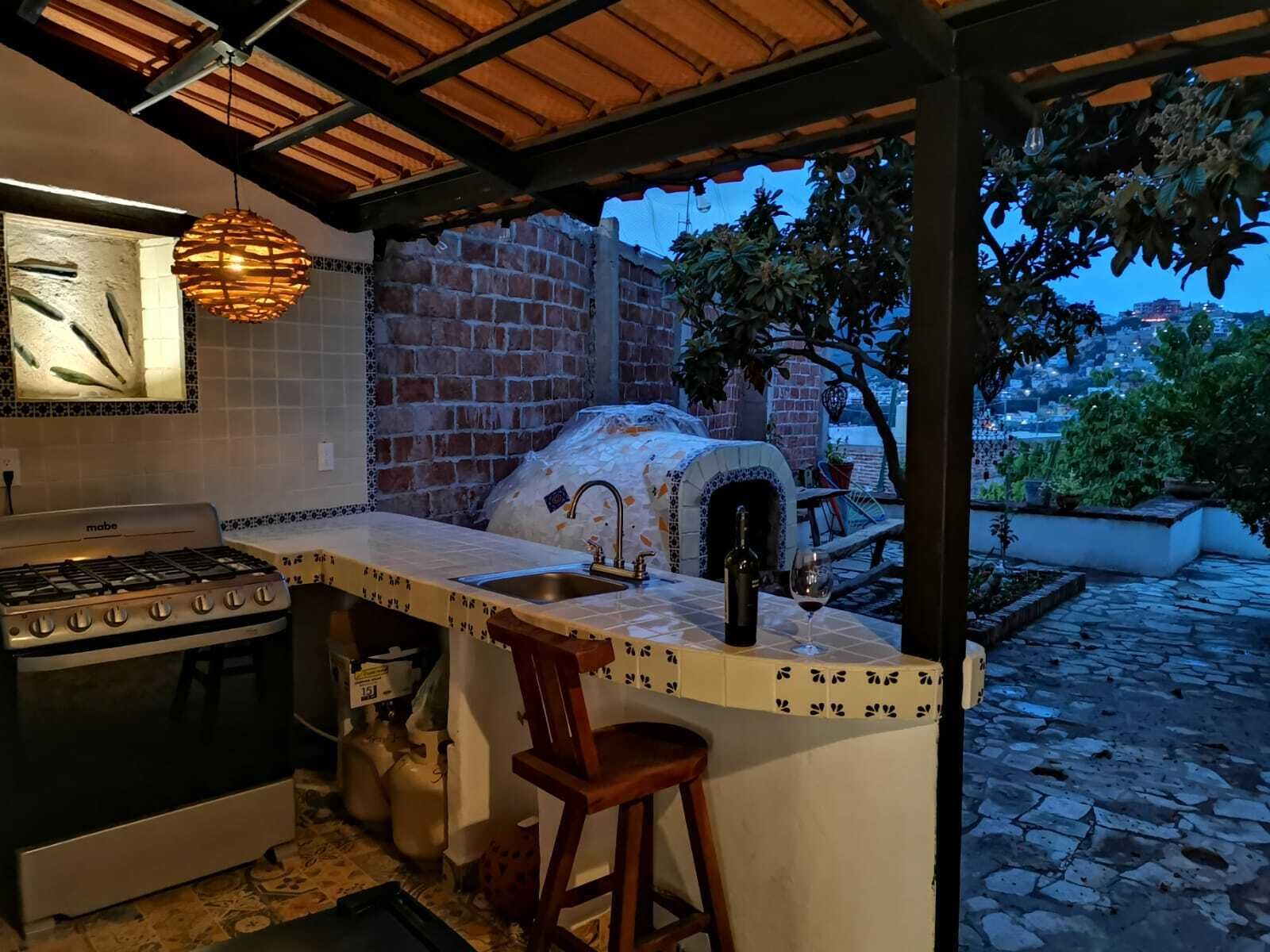 Une cuisine extérieure dotée d’un mur en briques, d’un four à pizza, d’une cuisinière à gaz et d’un coin bar avec des tabourets.