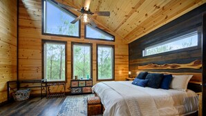 Un dormitorio de cabaña de madera con cama, ventanas grandes, ventilador de techo y diseño rústico en la pared.