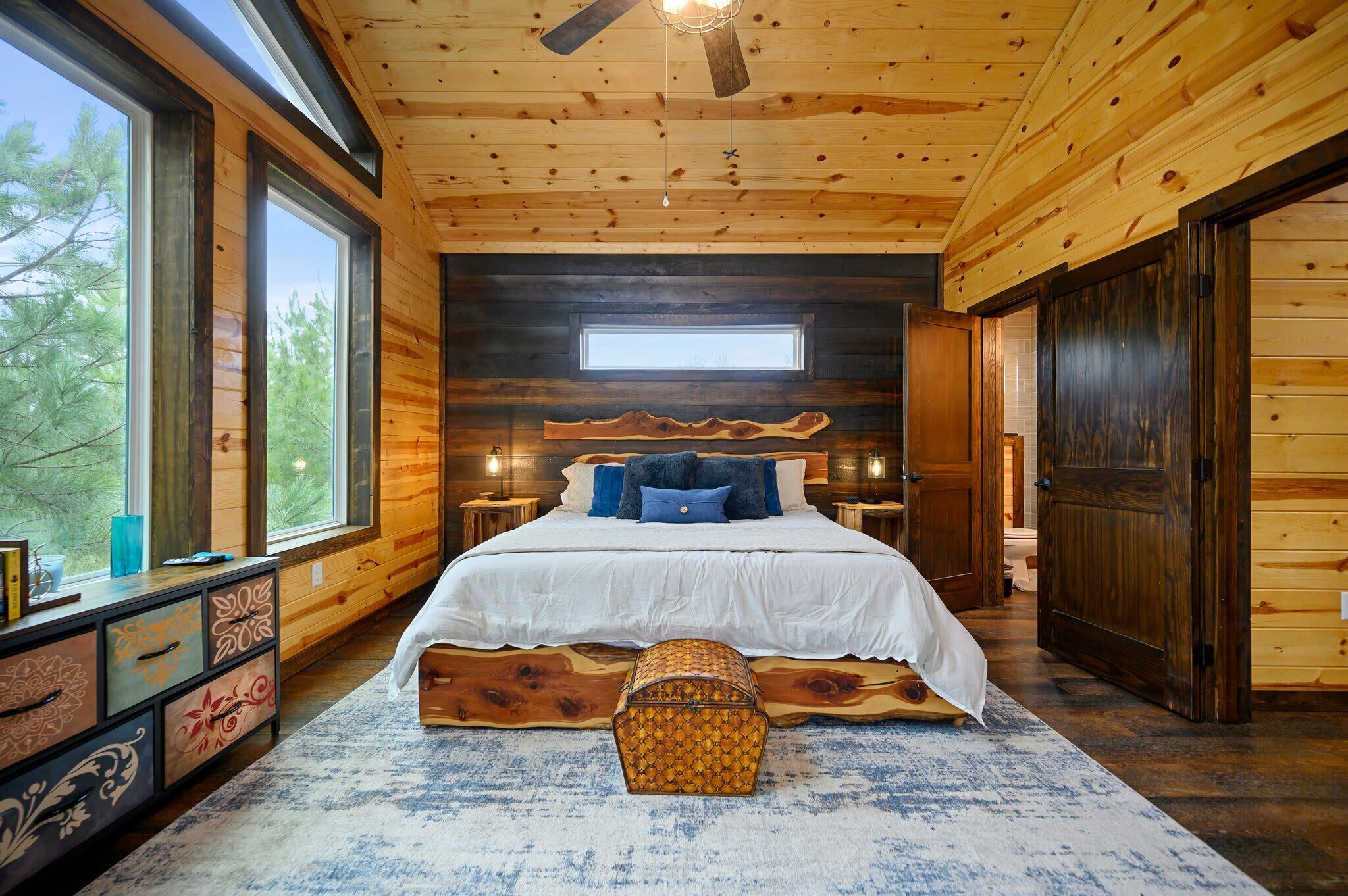Un dormitorio tipo cabaña de troncos con una cama grande, cabecera de madera y vistas a árboles a través de amplios ventanales.
