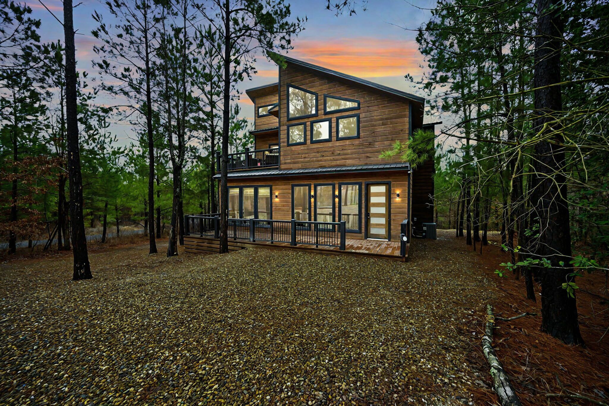 A modern two-story cabin with large windows and a balcony, surrounded by a forest.