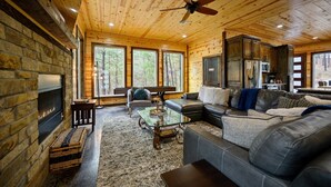 Interior acogedor de una cabaña con chimenea de piedra, sofá de cuero y un ventanal con vistas a un bosque.