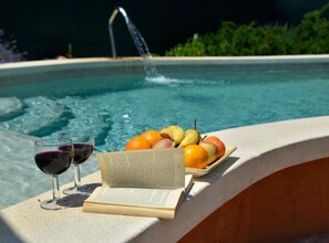 Escena junto a la piscina con un libro, copas de vino tinto y un plato de fruta.