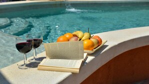 Un cadre agréable au bord de la piscine, avec un livre, des verres de vin rouge et une assiette de fruits.
