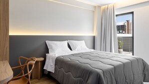 A modern hotel room with a large bed, a wooden side table, and a view of the city through a window. - Villano Hotel (Blumenau)
