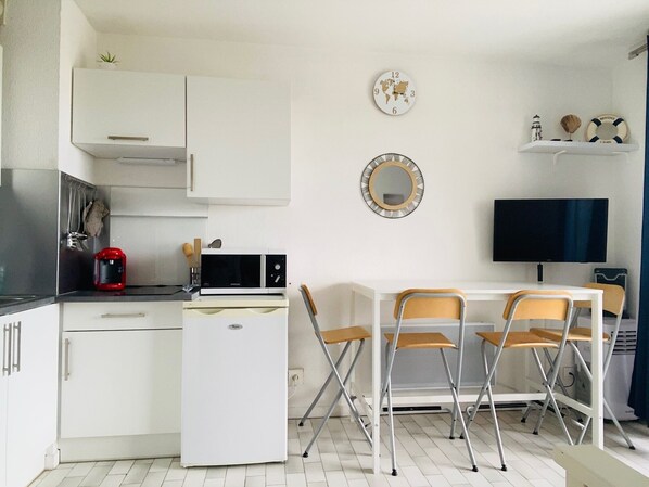 Een kleine keuken met witte kastjes, een magnetron, een rode koffiezetapparaat, en een eethoek met een tafel en stoelen.
