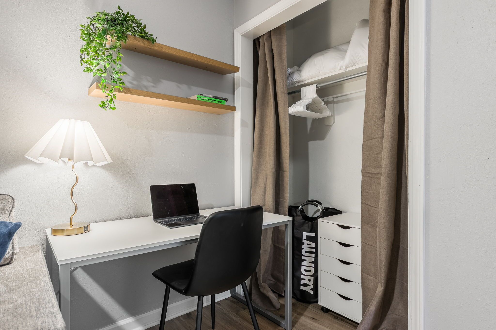 A bedroom with a white desk, a laptop, a black chair, a bedside lamp, a hanging plant, a closet with a 'laundry' bag, and a window with curtains.
