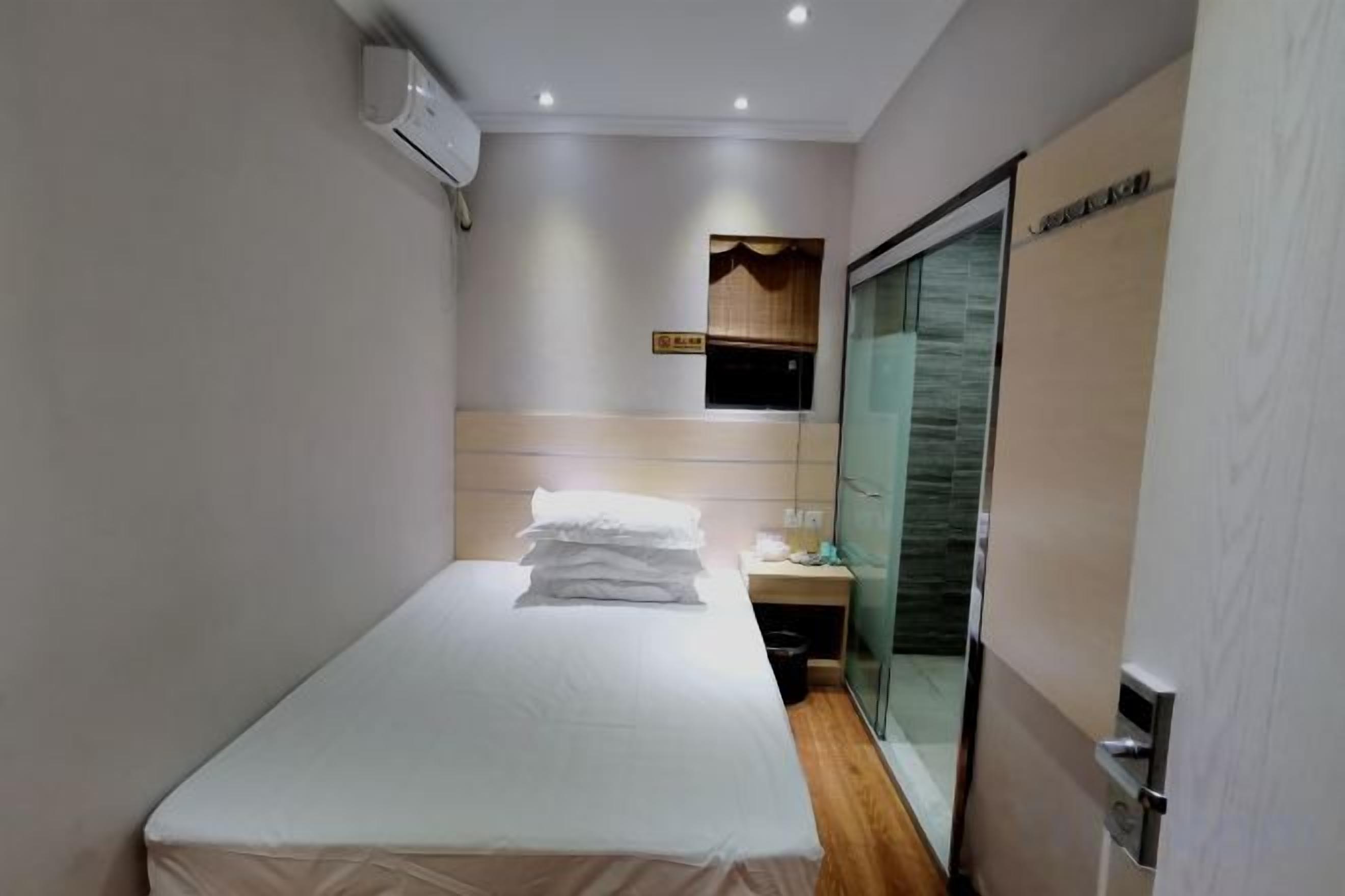 A narrow hotel room with a bed, a small table, and a glass door leading to a bathroom.